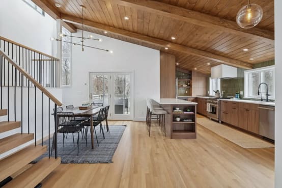 Open oak floating stair with black balusters beside a dining area and walnut kitchen under a wood plank ceiling.