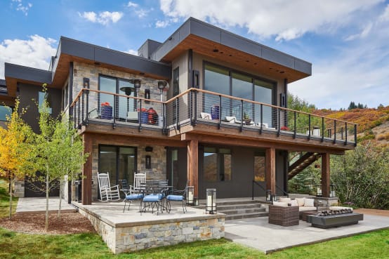 Cable railing with wood cap wraps the upper deck of a stone-and-timber modern home above a furnished patio.
