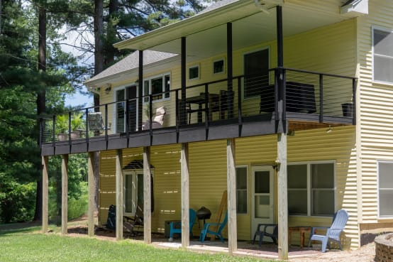 Black side-mounted cable railing on an elevated deck of a yellow home among pines.