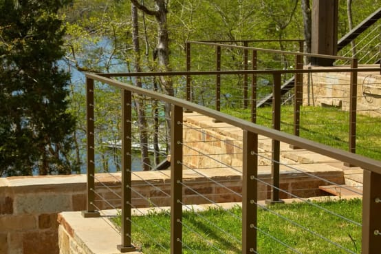 Bronze-finish posts and top rail with cable infill along a stone retaining wall above a lawn and lake.