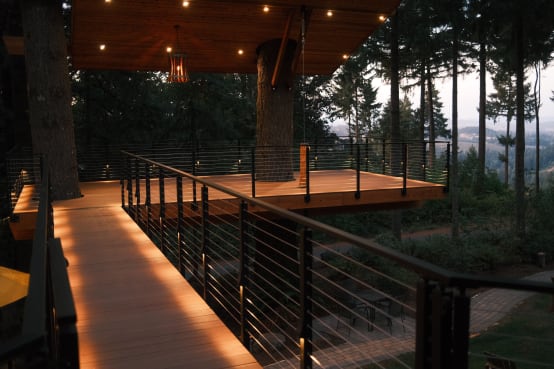 A glowing walkway lined with black cable railing leads to a covered treehouse deck built around a fir trunk.