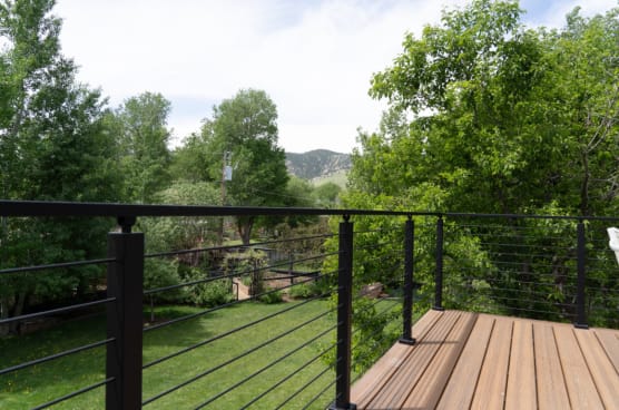 Black posts and top rail with cable infill along a wood deck overlooking a green lawn and trees.