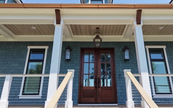 Front porch of a blue-shingled home with mahogany doors flanked by white posts and vertical cable railing.