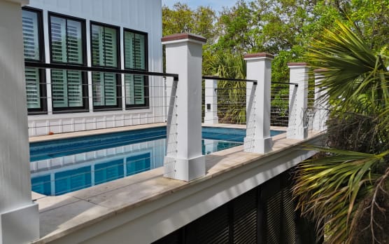 Black cable railing between white columns lines a raised pool deck among palms.