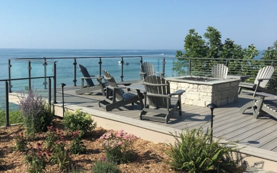 Glass panels between black posts around a composite deck with fire pit and Adirondack chairs facing the lake.