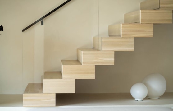 Side view of light oak stacked box treads climbing a cream wall with a slim black wall-mounted handrail.