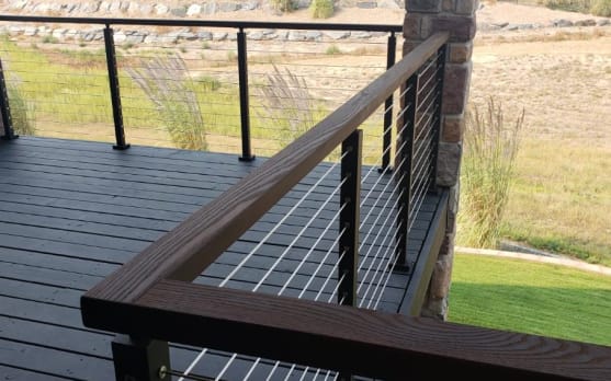 Black cable railing with a stained wood cap rail around a dark deck overlooking open meadow.