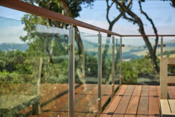 Glass panels between stainless posts with a wood cap rail edge a hardwood deck under oak trees.