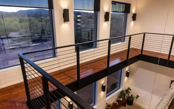 Black rod railing lines a wood-floored bridge spanning a double-height space with tall windows and sconces.