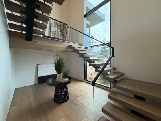 Black mono-stringer stair with thick oak treads and glass railing turns past a tall window.