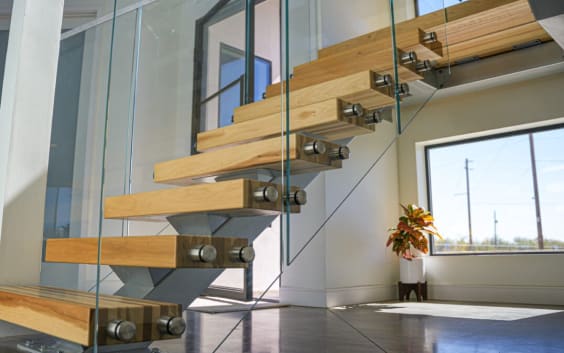 Light wood treads on a mono stringer with a frameless glass railing on standoff pins beside a tall window.