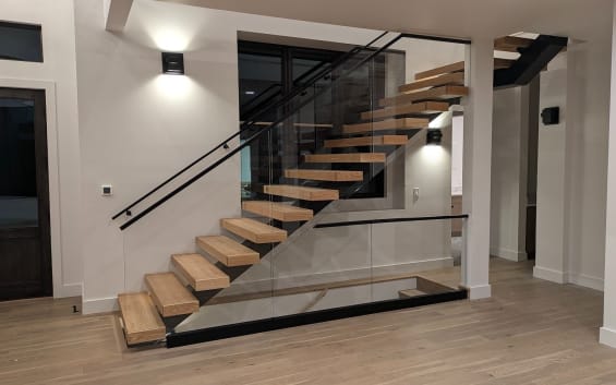 White oak floating treads on a black mono stringer turning at a landing with a frameless glass guard and wall handrail in a bright modern entry.