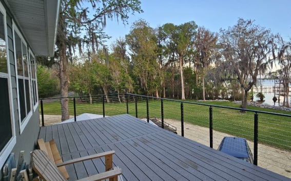 Black side-mounted cable railing borders a gray deck overlooking a lawn and cypress-lined lake.