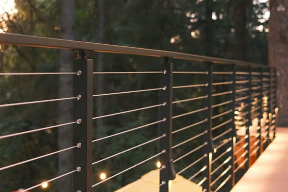 Black cable railing with bronze top rail and small post lights along an elevated forest walkway.