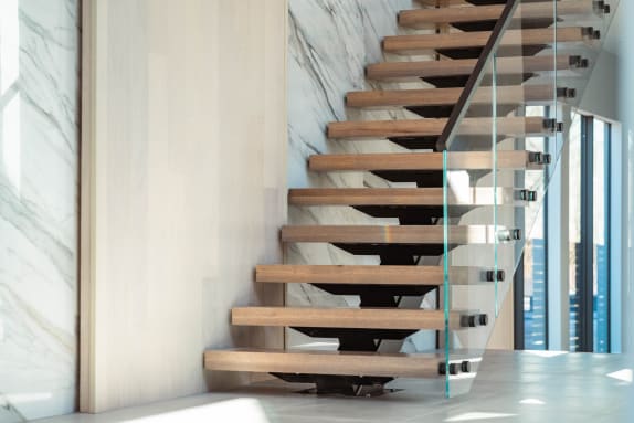 Oak treads on a black mono stringer with glass on black standoff pins beside a marble-clad wall.