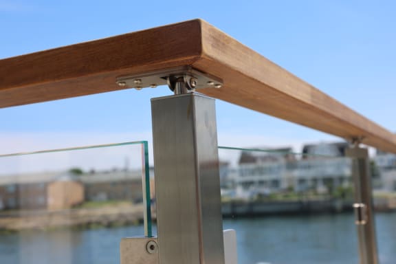 Close detail of a brushed stainless post supporting a hardwood top rail over glass panels with a waterfront behind.