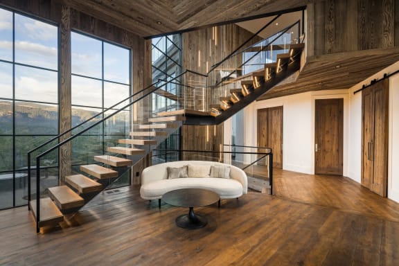 Wood floating treads with LED accents on black stringers switch back beside floor-to-ceiling windows, with glass railing and black handrail in a reclaimed-wood interior.