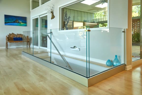 Frameless glass panels in a brushed base rail enclose a stair opening on a maple floor with a stainless handrail.