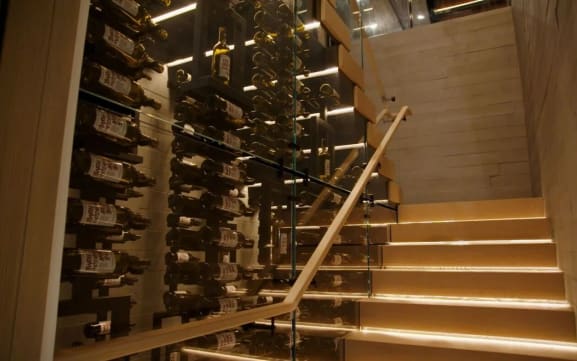 LED-lit light wood stack stair with glass railing and wood handrail running beside a glass-walled wine cellar.