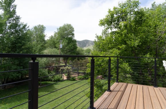 Black posts and top rail with cable infill along a wood deck overlooking a green lawn and trees.