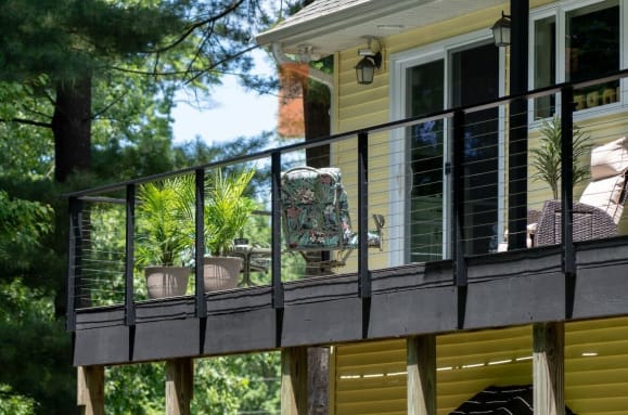 Black slim side-mount posts with cable infill and beverage handrail along a raised deck beside a yellow house.