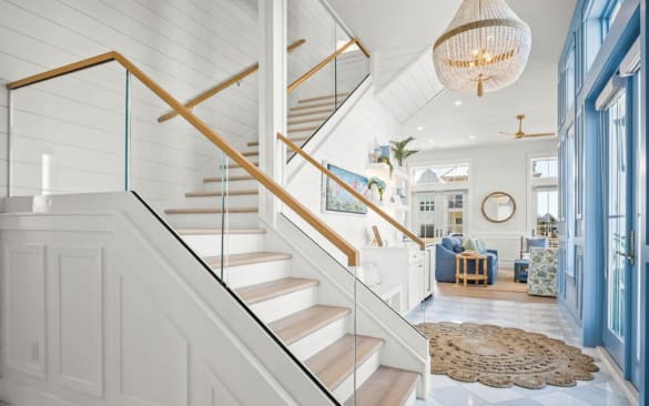 Frameless glass in a white base rail with a white oak handrail lines a shiplap stair in a bright coastal living room.