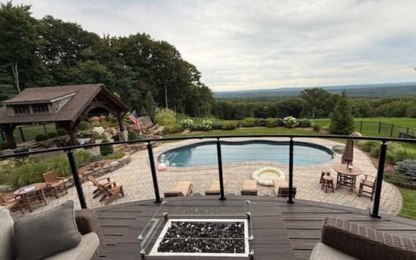 Black cable railing curves around a dark composite deck with a fire table overlooking a pool and wooded valley.