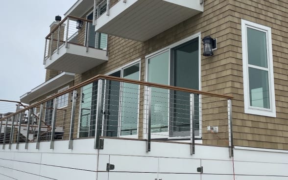 Stainless side-mount posts with cable infill and wood cap on decks of a shingled coastal home.