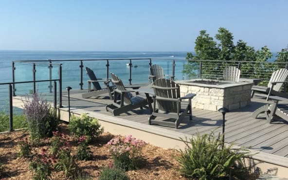 Glass panels between black posts around a composite deck with fire pit and Adirondack chairs facing the lake.