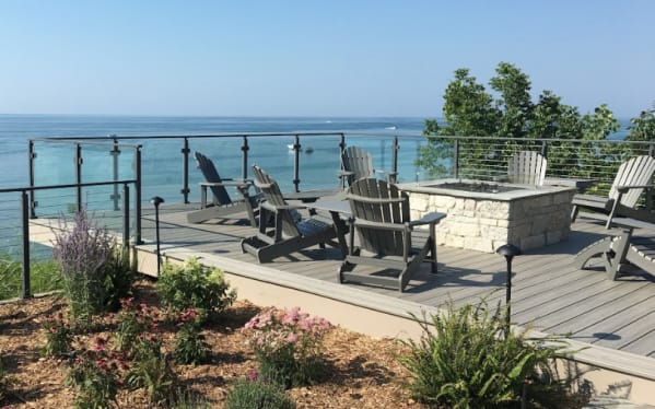 Glass panels between black posts around a composite deck with fire pit and Adirondack chairs facing the lake.