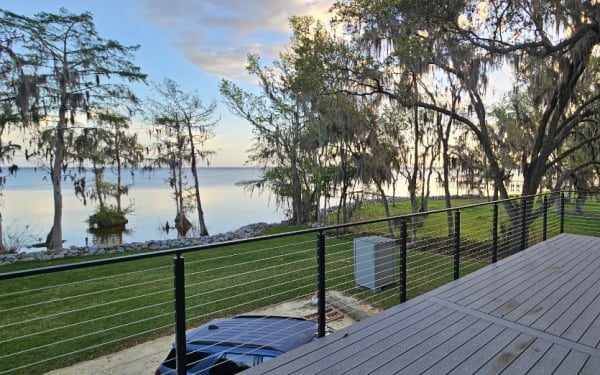 Black cable railing along a gray deck overlooking a lake framed by mossy oaks.