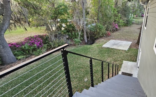 Black side-mount cable railing with a wood cap follows a gray exterior stair beside a green-sided house.