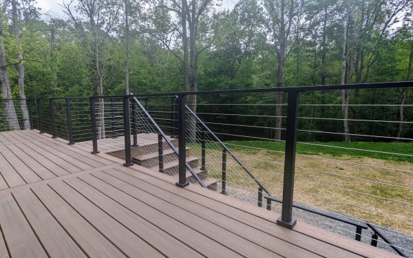 Black cable railing along a brown composite deck and its short stair down to a lawn edged by trees.