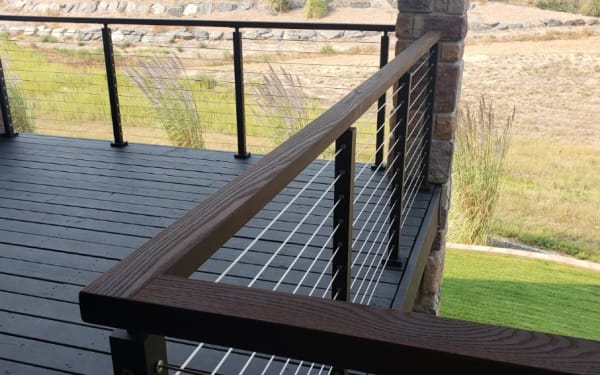 Black cable railing with a stained wood cap rail around a dark deck overlooking open meadow.