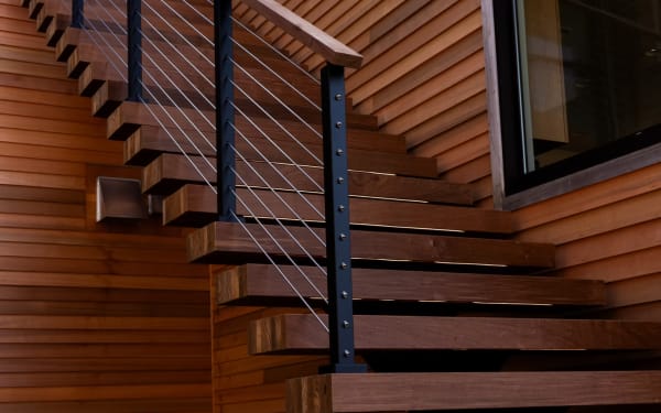 Thick Ipe floating treads with black-post cable railing against warm horizontal wood paneling.