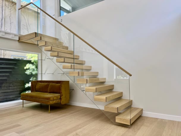 Thick white oak treads cantilever from a white wall beside full-height frameless glass and a slim wood handrail, with an ochre loveseat below.