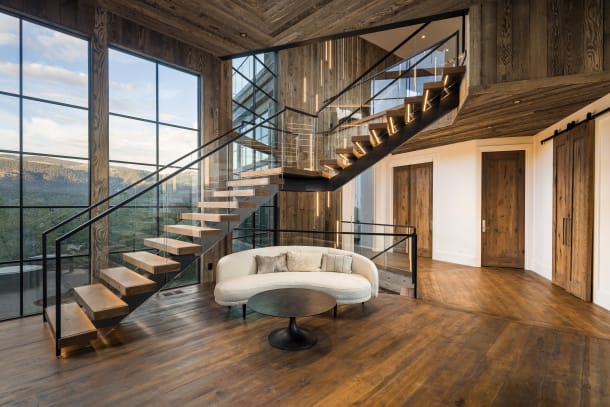 Wood floating treads with LED accents on black stringers switch back beside floor-to-ceiling windows, with glass railing and black handrail in a reclaimed-wood interior.