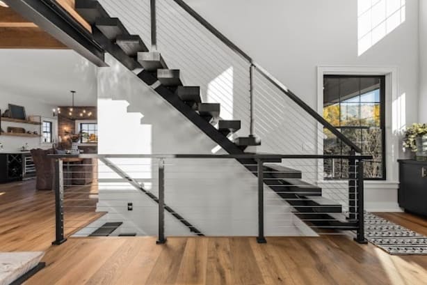 Black floating treads on a mono stringer with cable railing rise past a white shiplap wall above a wood floor.