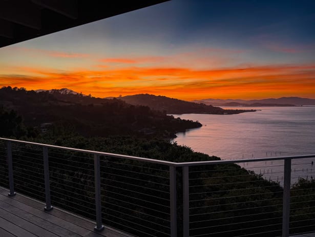 Composite deck with dark cable railing framing a vivid orange sunset over hills and water.