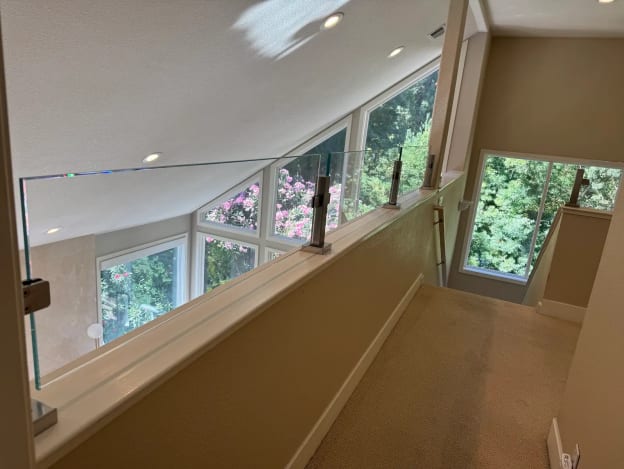 Stainless square posts hold glass panels along a loft knee wall beside angled windows and greenery.