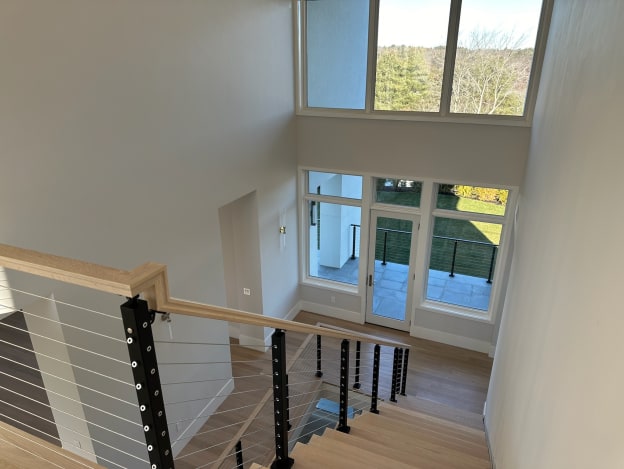 Black-post cable railing with oak handrail looks down a white oak stair to a window-filled entry.