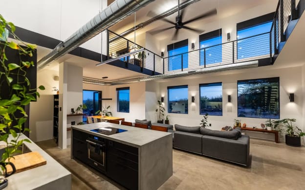 Black rod railing runs along a loft above a concrete kitchen island and living area.