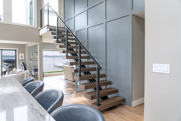 Open wood treads on a black stringer with black post cable railing beside a gray paneled wall in a lake house.