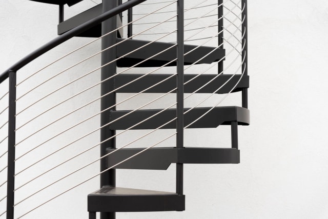 Black steel spiral stair with metal treads and cable railing against a white wall.