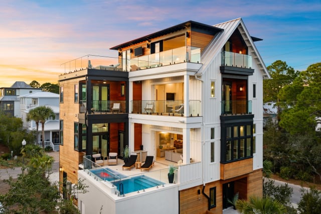 Multi-story coastal home at dusk with frameless glass railings on every balcony, rooftop deck and elevated pool.