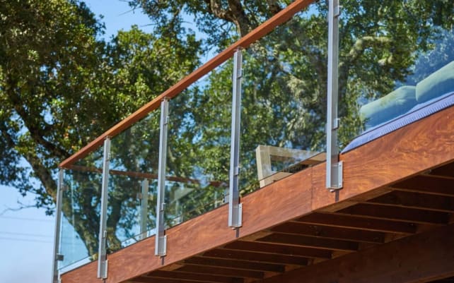 Stainless side-mount posts hold glass panels under a wood top rail on a redwood deck among oak trees.