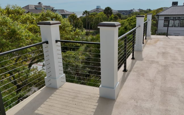 White columns and black cable railing at the corner of an upper deck surrounded by trees.