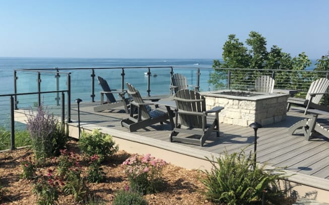 Glass panels between black posts around a composite deck with fire pit and Adirondack chairs facing the lake.