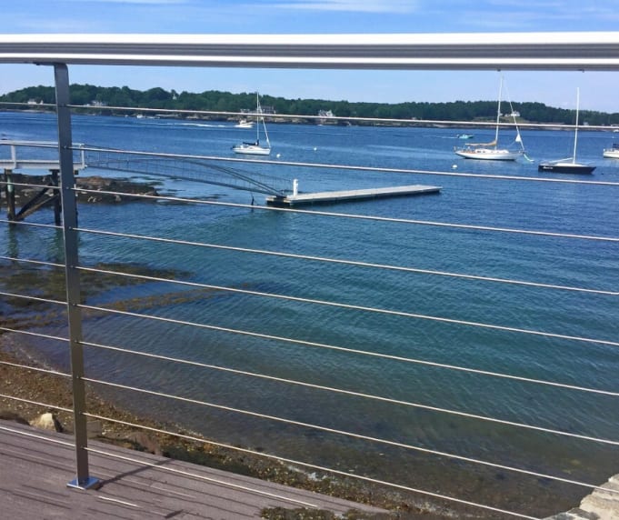 Stainless posts with round handrail and cable infill frame a harbor view with moored sailboats.