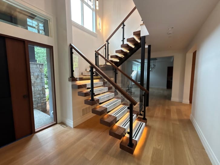 Walnut floating treads glow with LED lighting along a mono stringer with glass panels and a wood handrail in a bright foyer.